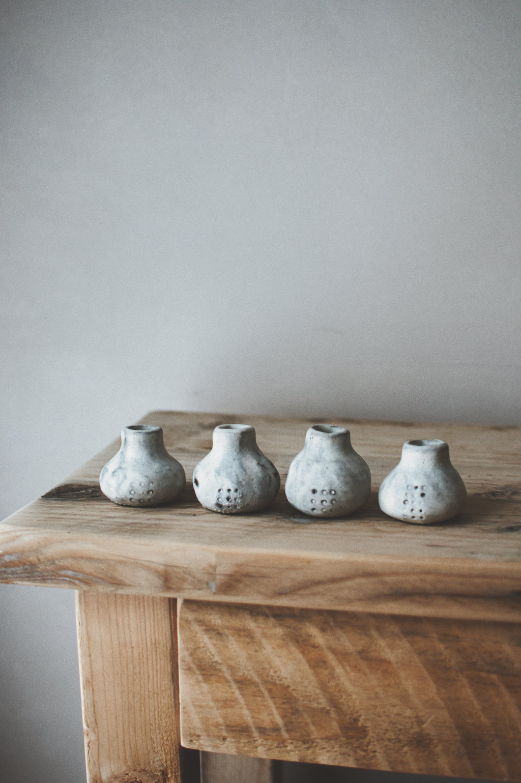 Four small handmade ceramic vases with a textured, speckled gray and white glaze, lined up on a rustic wooden table against a neutral gray wall.