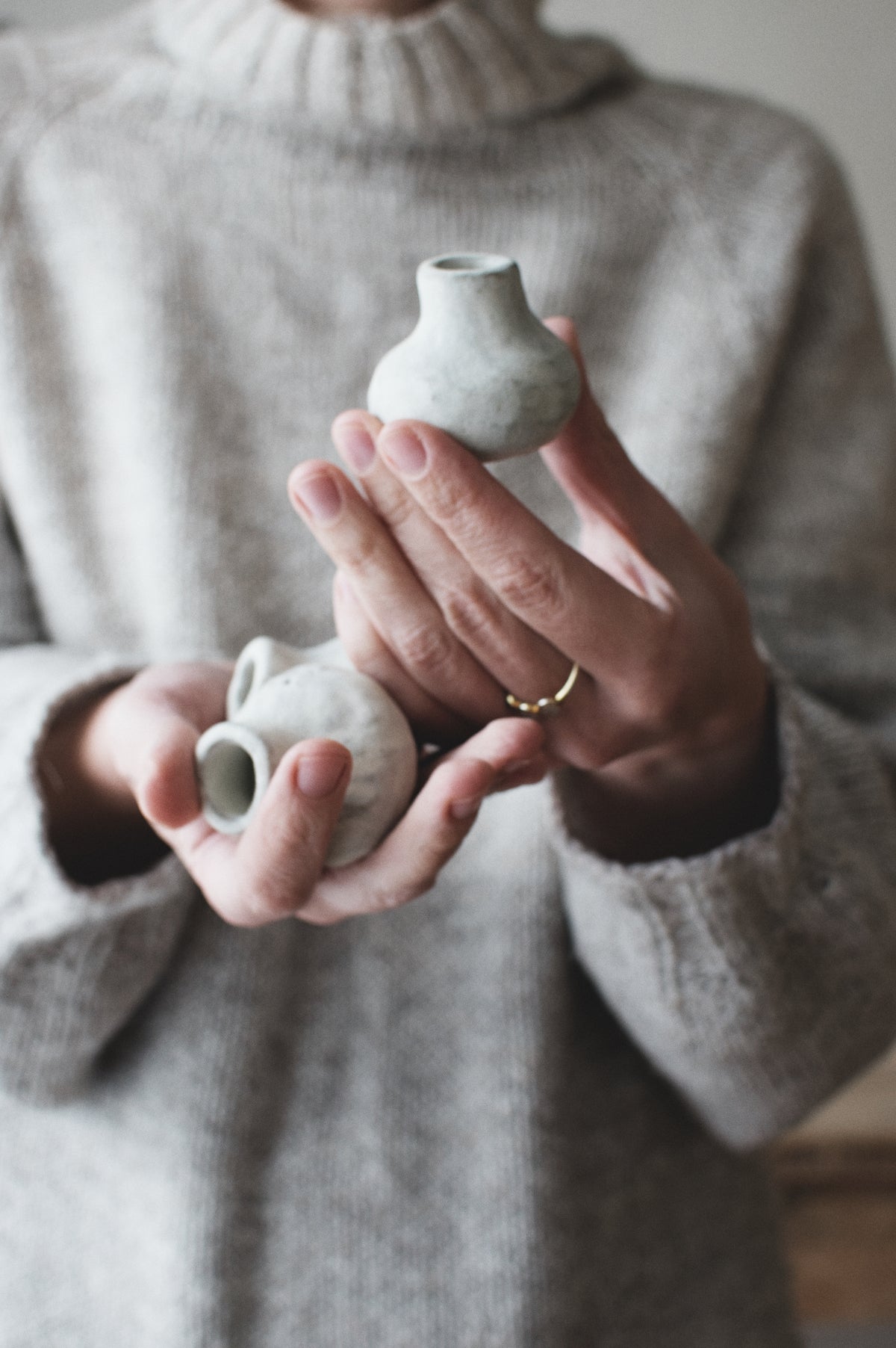 A person in a cozy gray knit sweater holds two small handmade ceramic vases with a matte, textured off-white finish. The person is wearing a gold ring on one finger, and the background is softly blurred.